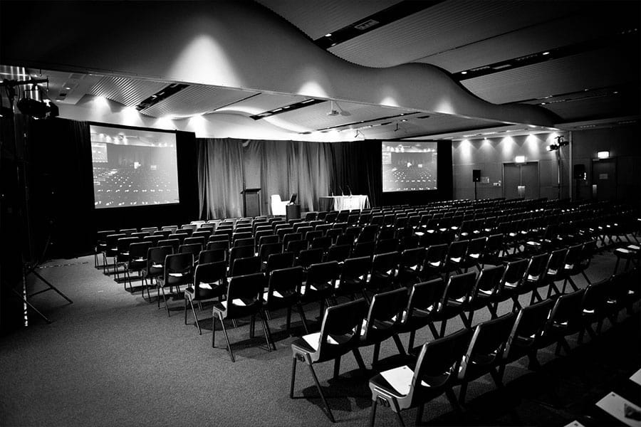 A black and white image of a large room in the conference centre set up for an event with two large projector screens and rows of seats.